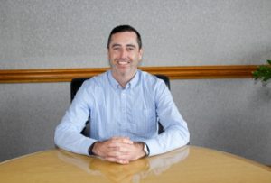 a man in a blue shirt sits at a table with his hands folded while smiling